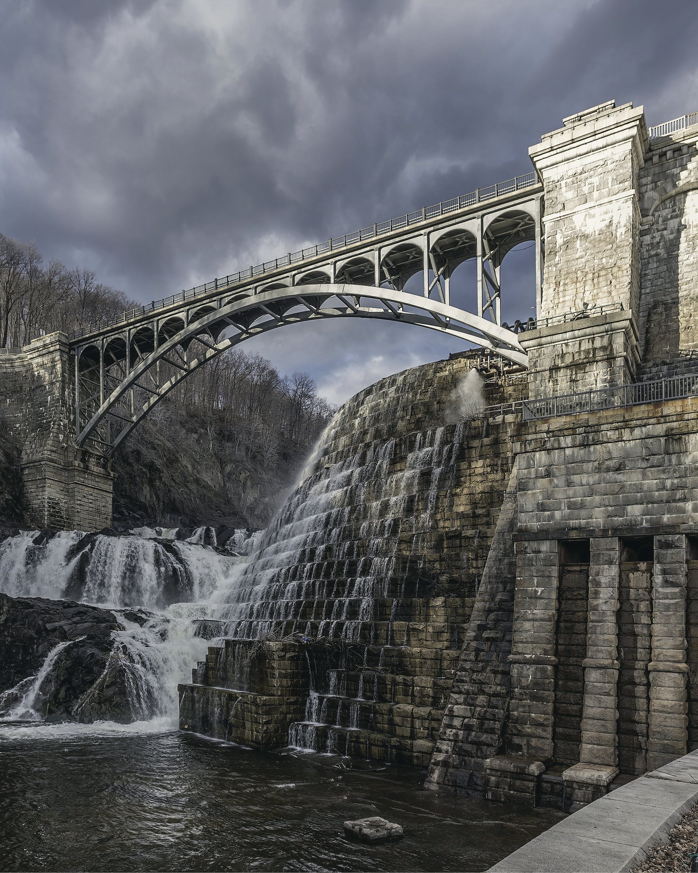 The New Croton Dam under a storm sky: the steel arch bridge over the spillway, water falling down the stepped stone face.